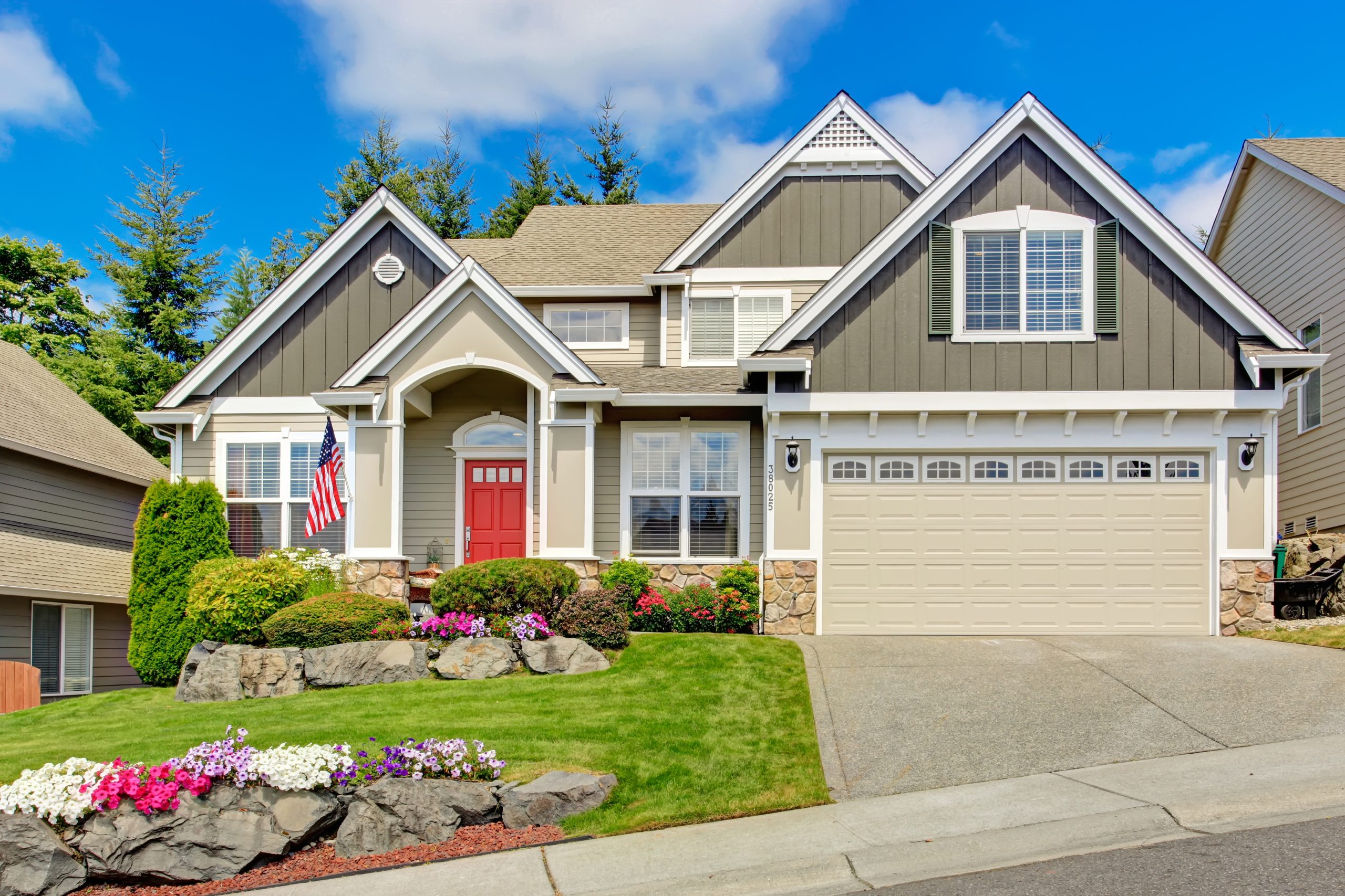 Grey house exterior with entrance porch and red door. Beautiful front yard landscape with vivid flower and stones